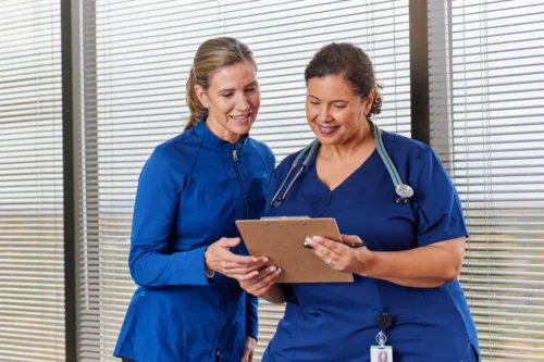 Two healthcare professionals in blue uniforms review information together on a clipboard while standing in front of hospital windows.